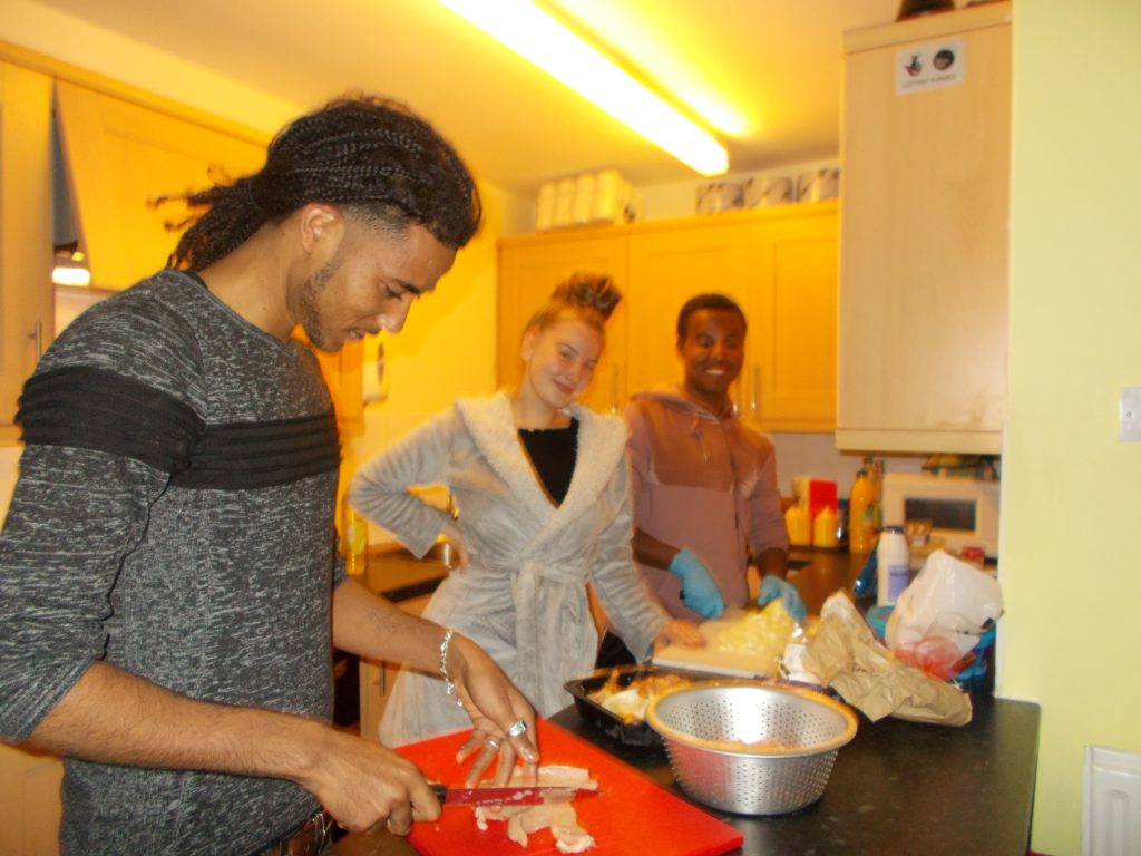 3 young people cooking, 1 man is in the foreground cooking chicken, the other 2 are behind a counter posing and smiling